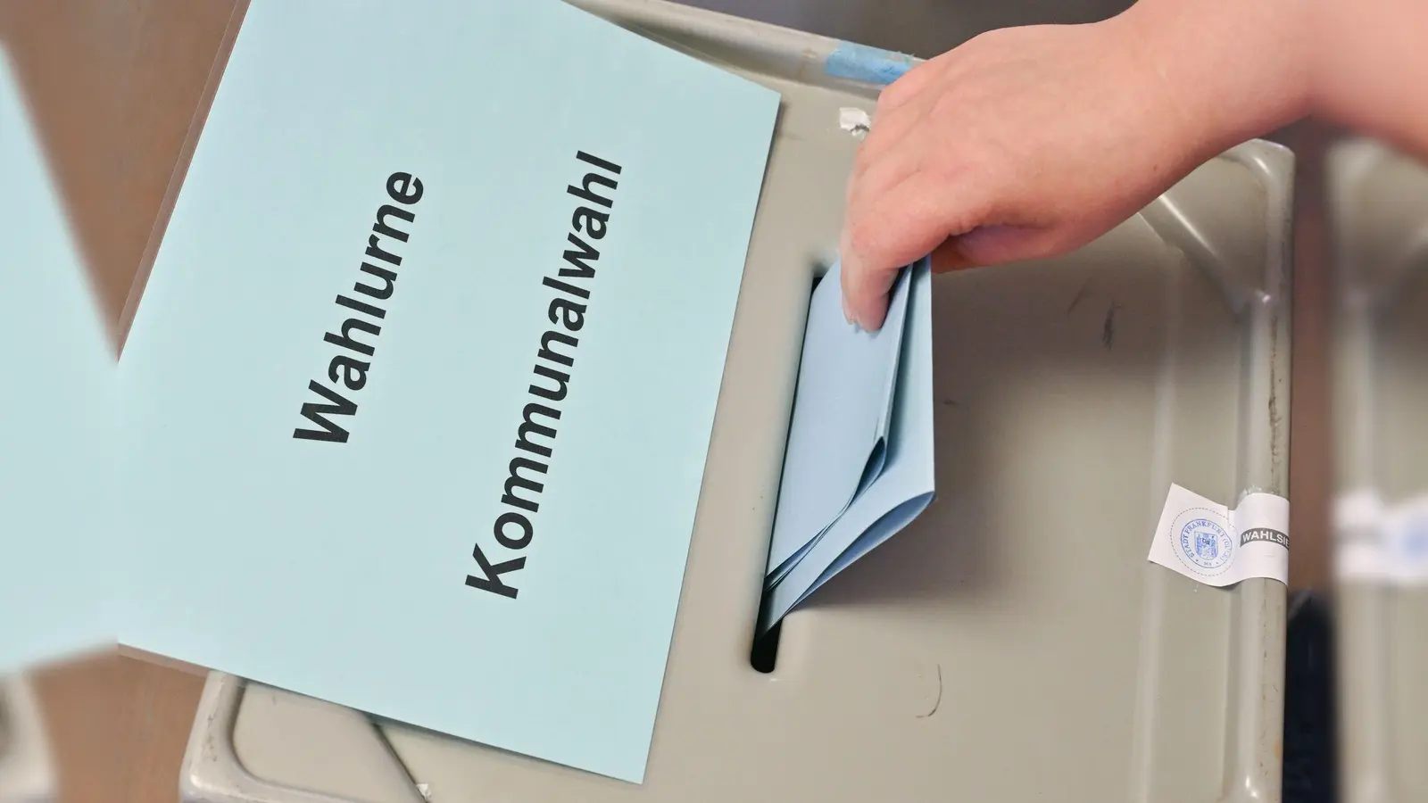 Eine Hand steckt einen Stimmzettel in eine Wahlurne mit der Aufschrift „Wahlurne Kommunalwahl“. (Symbolbild: Patrick Pleul/dpa)